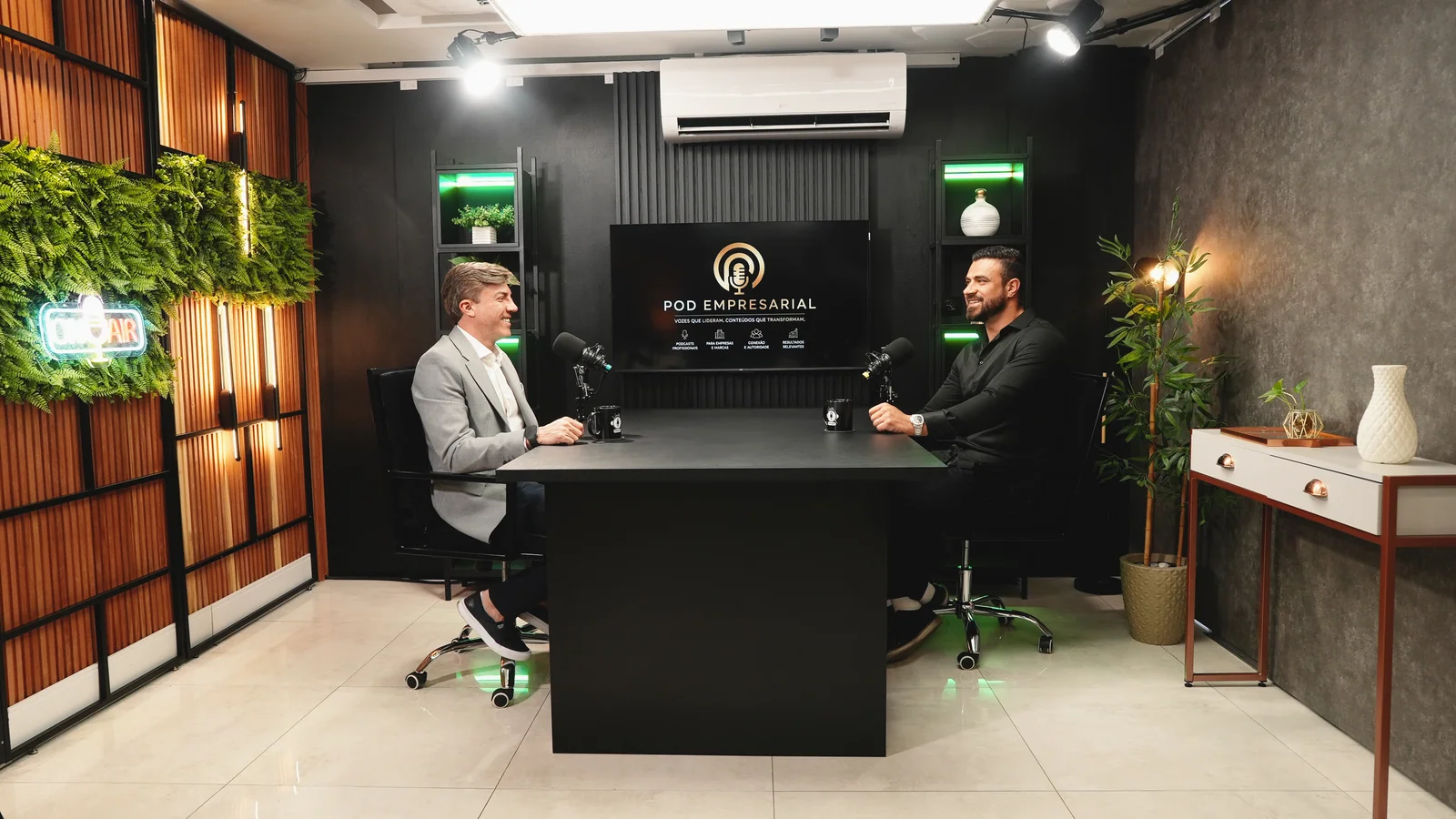 Mesacast corporativo do Estúdio Master com mesa preta, TV exibindo a vinheta do programa, iluminação RGB e letreiro neon On Air